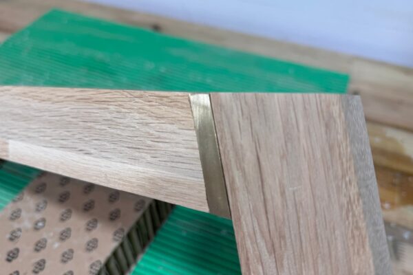 Primer plano de la unión entre dos piezas de roble con un inserto de latón en la estructura de una mesa de centro artesanal.
