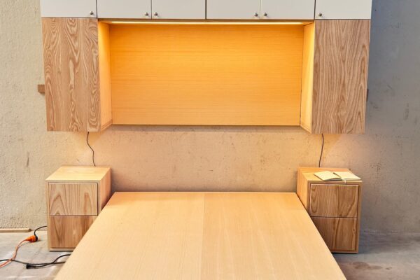 Dormitorio compacto con cama, mesillas y módulos superiores fabricados a medida en madera de fresno y acabado blanco mate con iluminación LED integrada.
